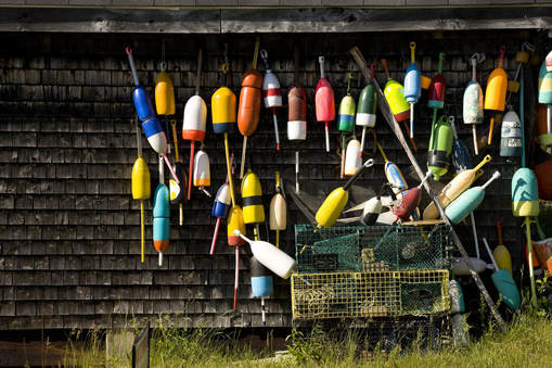 A photograph of many lobster buoys in various bright colors leaning against a wall with cedar shingles.