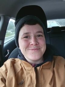 Photo of a smiling person with a lip piercing and wearing a black stocking cap and a tan jacket