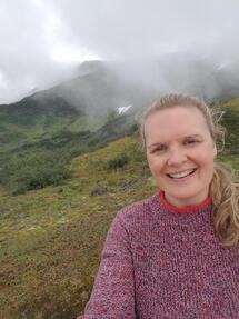 Photo of a woman wearing a pink sweater, smiling in front of a fog hazed mountain